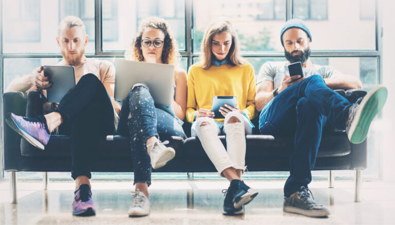 Four young professionals on a sofa, each using a laptop, tablet or phone