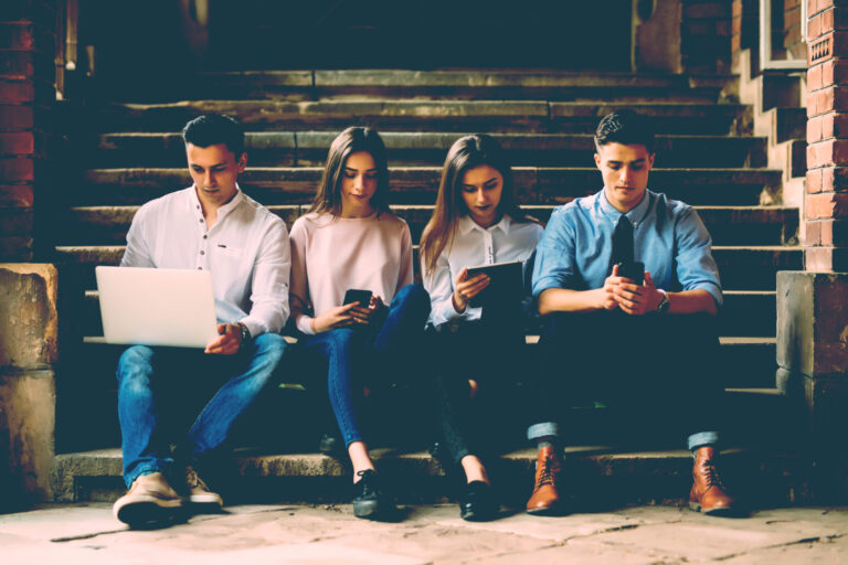 Four young professionals sitting on stone steps using a laptop, tablets and phones