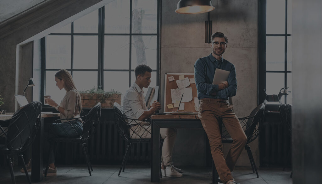 Small team working in a modern loft office, one man standing with a laptop