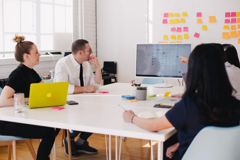 Team gathered around a desk discussing a design on a monitor, with sticky notes on the wall