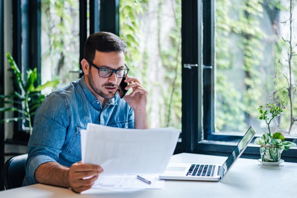 Corporate communication. Portrait of busy young male manager working remotely, holding paper and reading financial document, using laptop and talking on mobile phone, sitting at desk.