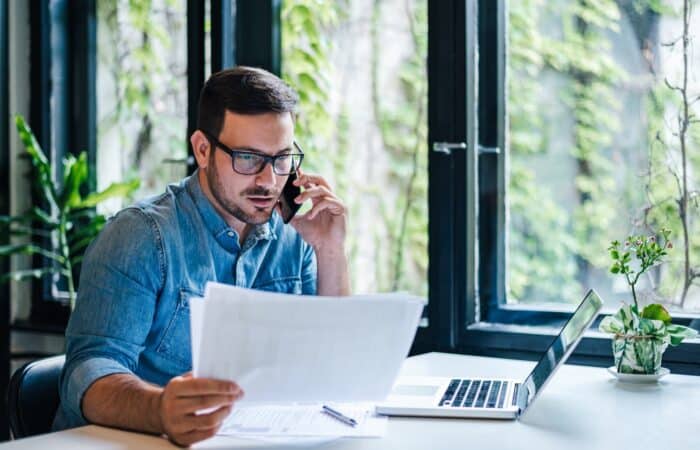 Corporate communication. Portrait of busy young male manager working remotely, holding paper and reading financial document, using laptop and talking on mobile phone, sitting at desk.